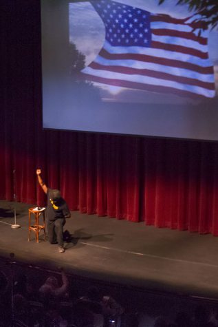 W. Kamau Bell kneels and holds up a black power salute while hosting the 26th annual Leonardo Dorantes Memorial Lecture.