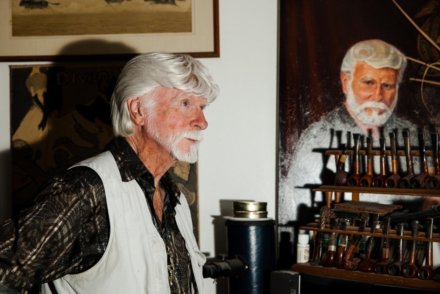 Ron Atwood stands in his home office with a portion of his antique tobacco pipe collection and a self portrait painting from 1998 done by a friend.