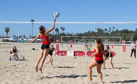 With a sign “Smiley Irey” in view, Irey Sandholdt (No. 12) jumps up to win a point in a match with teammate Paige Rudy (No. 9) against Moorpark College on Friday, April 21 at West Beach in Santa Barbara Calif.
