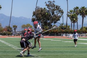 SBCC player Teagan Cleary (No. 21) engages in a physical confrontation while battling for possession during a men's lacrosse semi-final game at La Playa Stadium on April 19, in Santa Barbara, Calif. The image shows Cleary in mid-air as he collides with two of the three defenders near the center of the field.